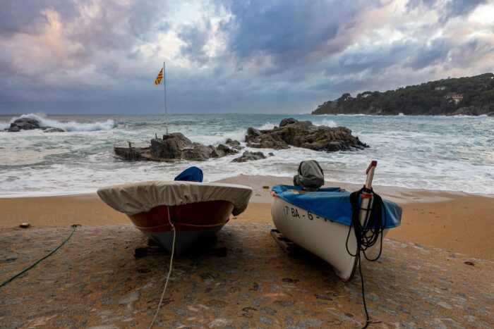 Two boats on a sandy beach in Calella de Palafrugell with waves and rocks in the background.